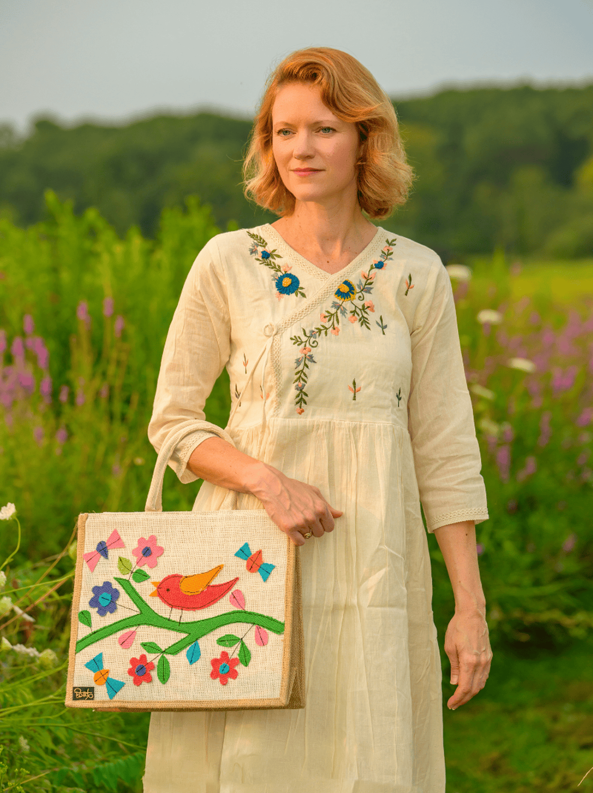 Woman in an embroidered dress holding a colorful tote bag in a field with flowers and trees.