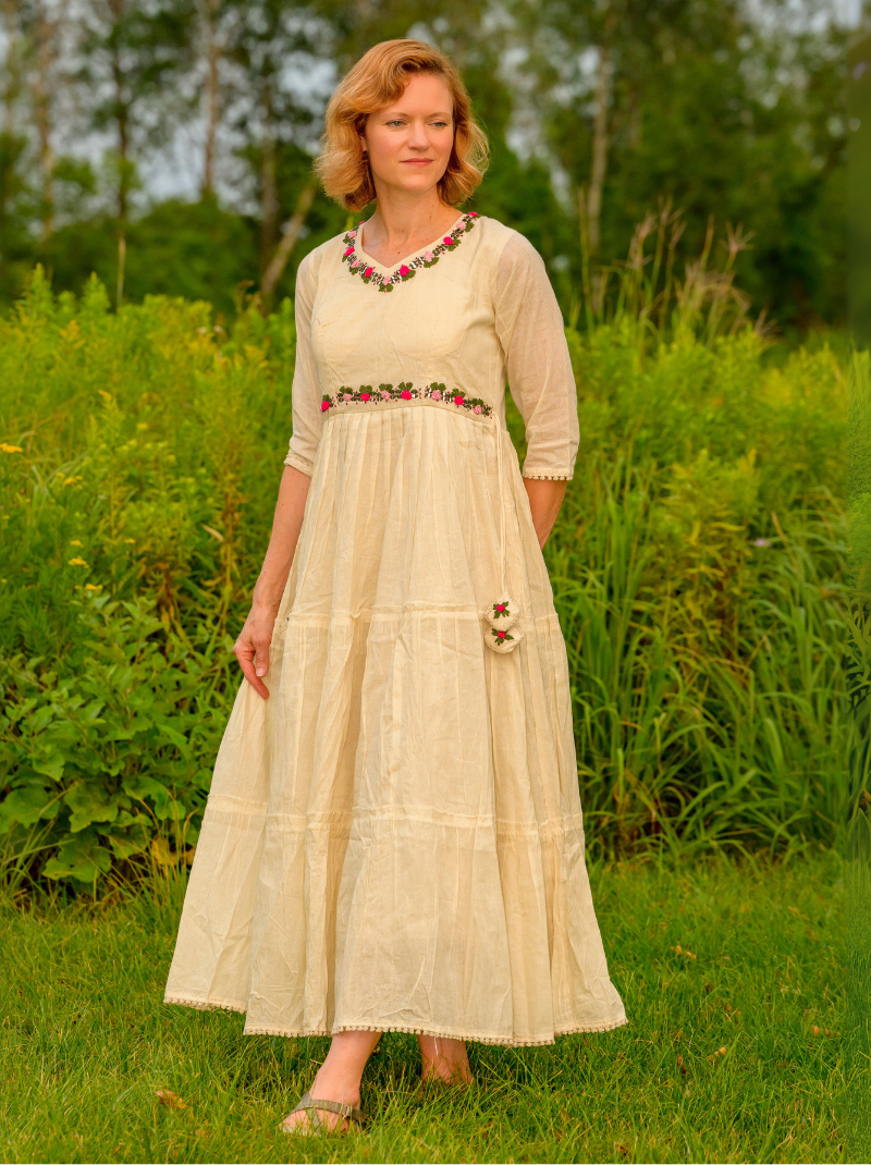 Woman in a long, light-colored dress with floral embellishments standing in a grassy field.