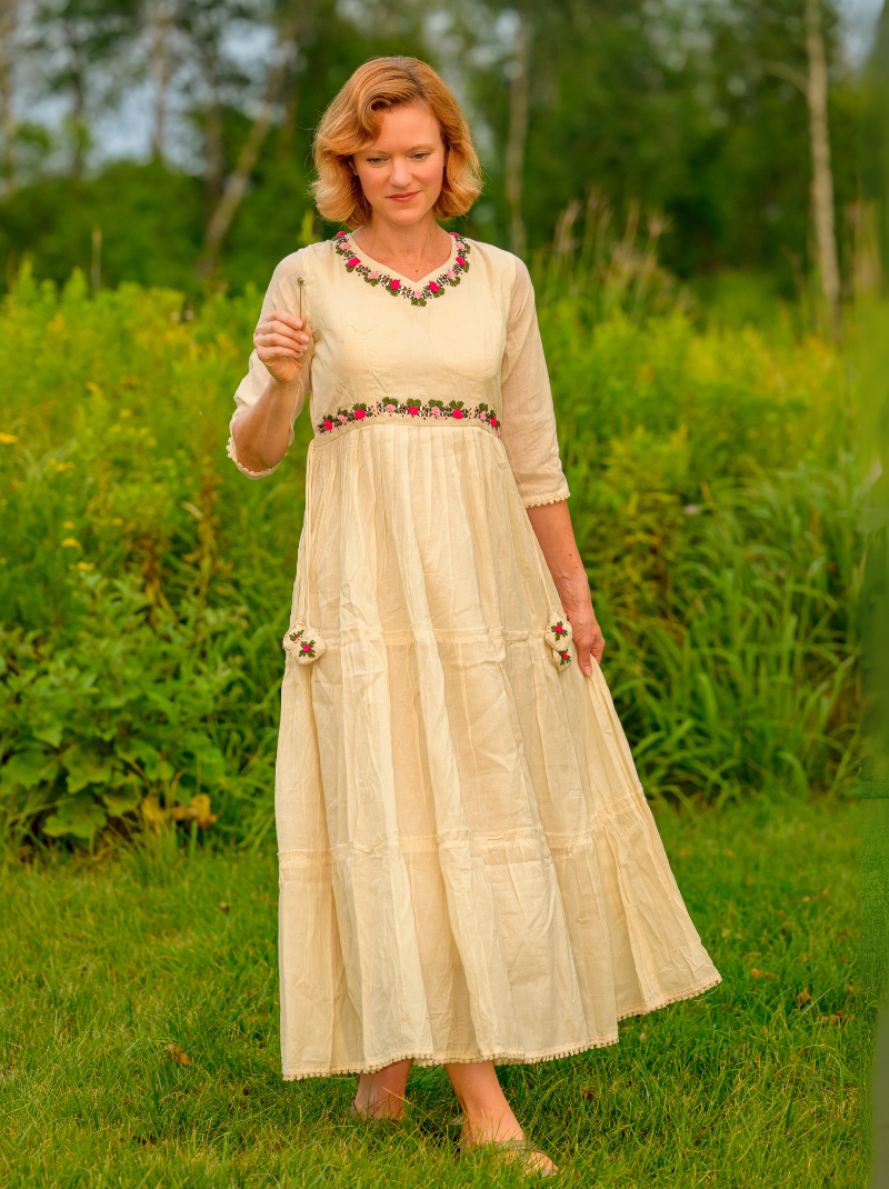Woman in a long, light-colored dress with floral embellishments standing in a grassy field.