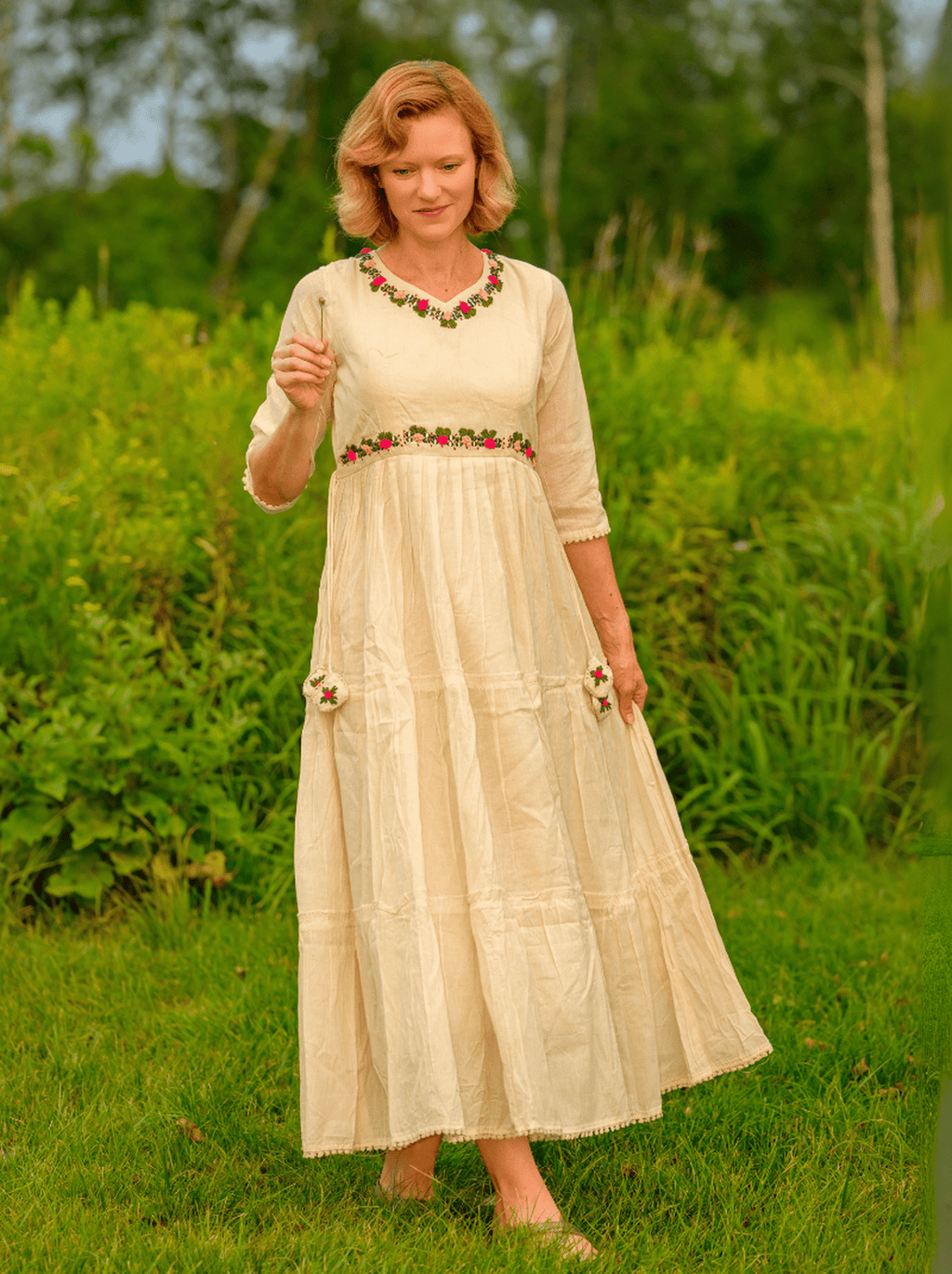 Woman in a long, light-colored dress with floral embellishments standing in a grassy field.