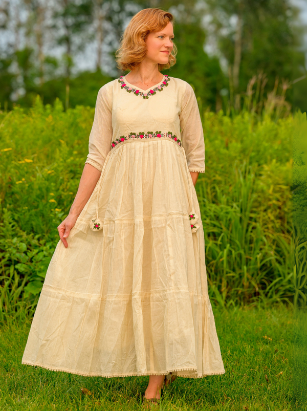 Woman in a long beige dress with floral embellishments standing in a grassy field.