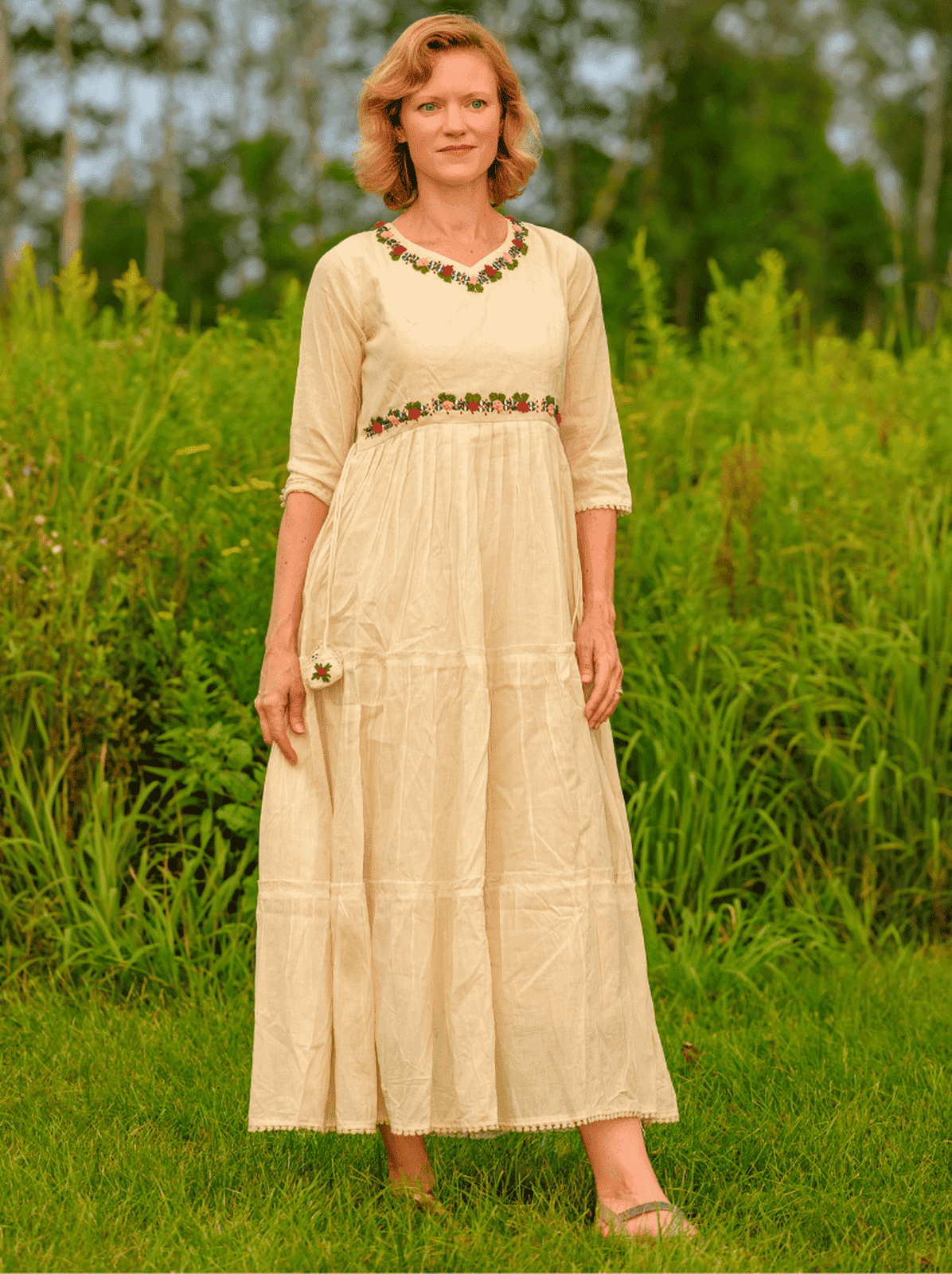 Woman wearing a long, light-colored dress with floral embroidery in a grassy field.
