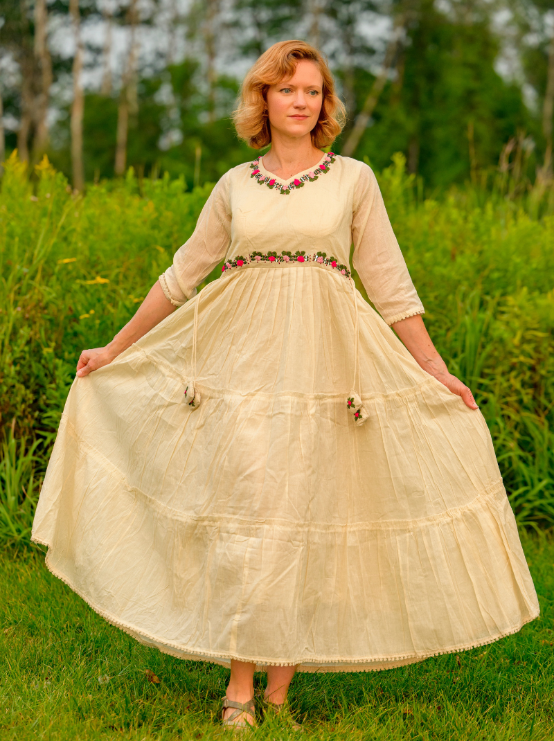 Woman wearing a long, light-colored dress with a floral neckline in a grassy outdoor setting.