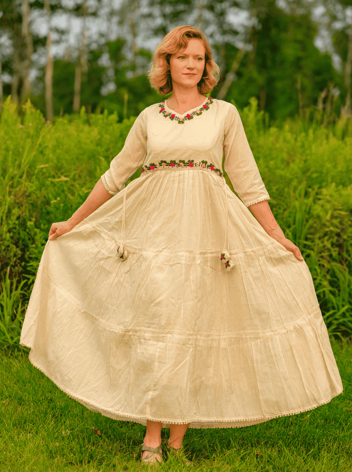 Woman wearing a long, light-colored dress with a floral neckline in a grassy outdoor setting.
