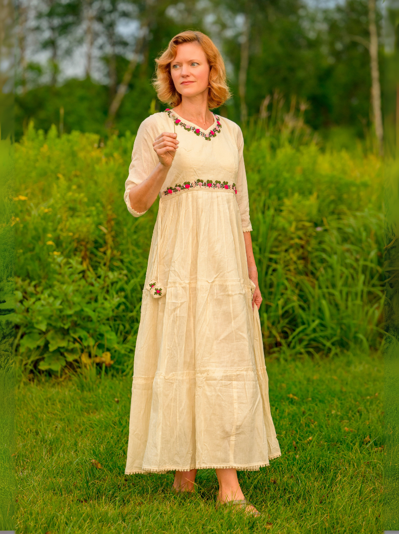 Woman in a long, light-colored dress with floral embroidery standing in a grassy field.