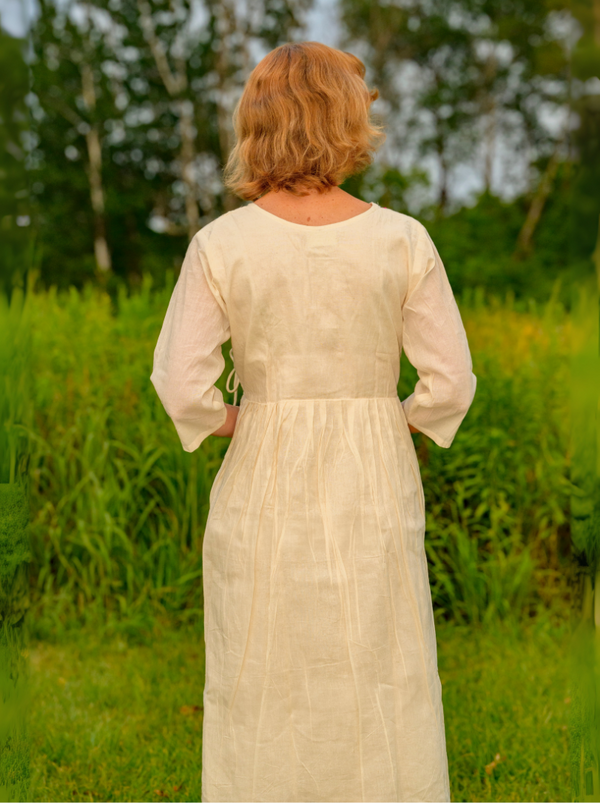 Woman in a long, light-colored dress standing in a grassy field with trees in the background.
