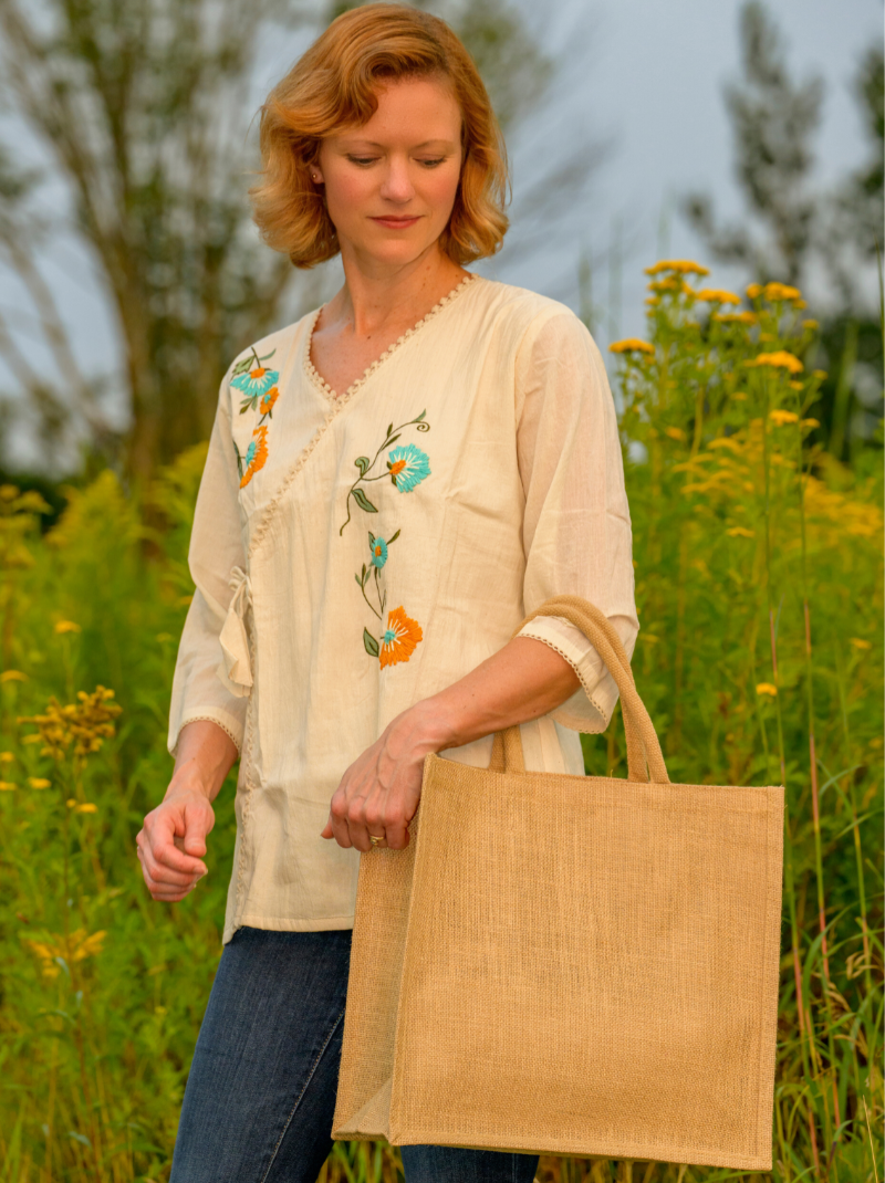 Woman holding a beige tote bag in a field of tall grass and wildflowers