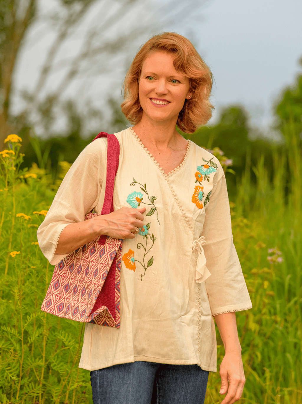 Woman standing in a field wearing a floral embroidered blouse and holding a pink bag.