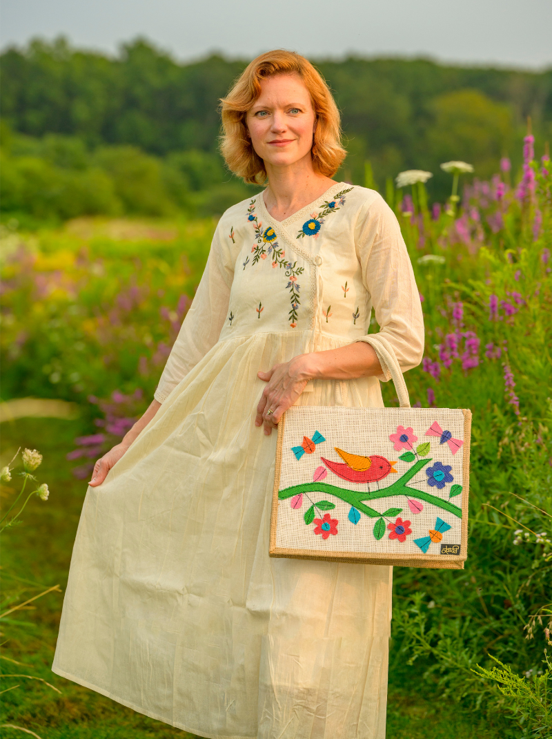 Woman in a white dress holding a colorful bag in a field with flowers and trees.
