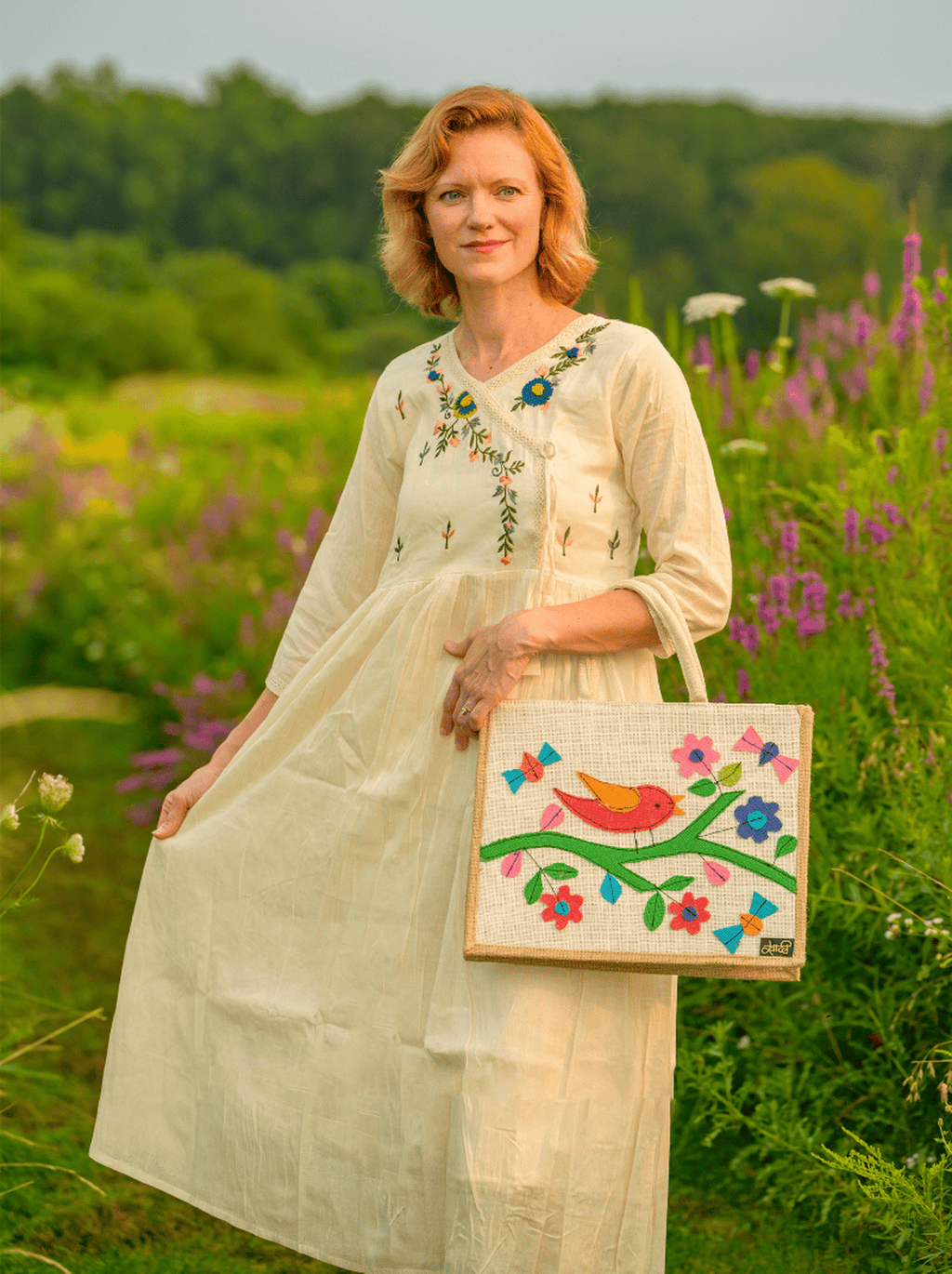 Woman in a white dress holding a colorful bag in a field with flowers and trees.
