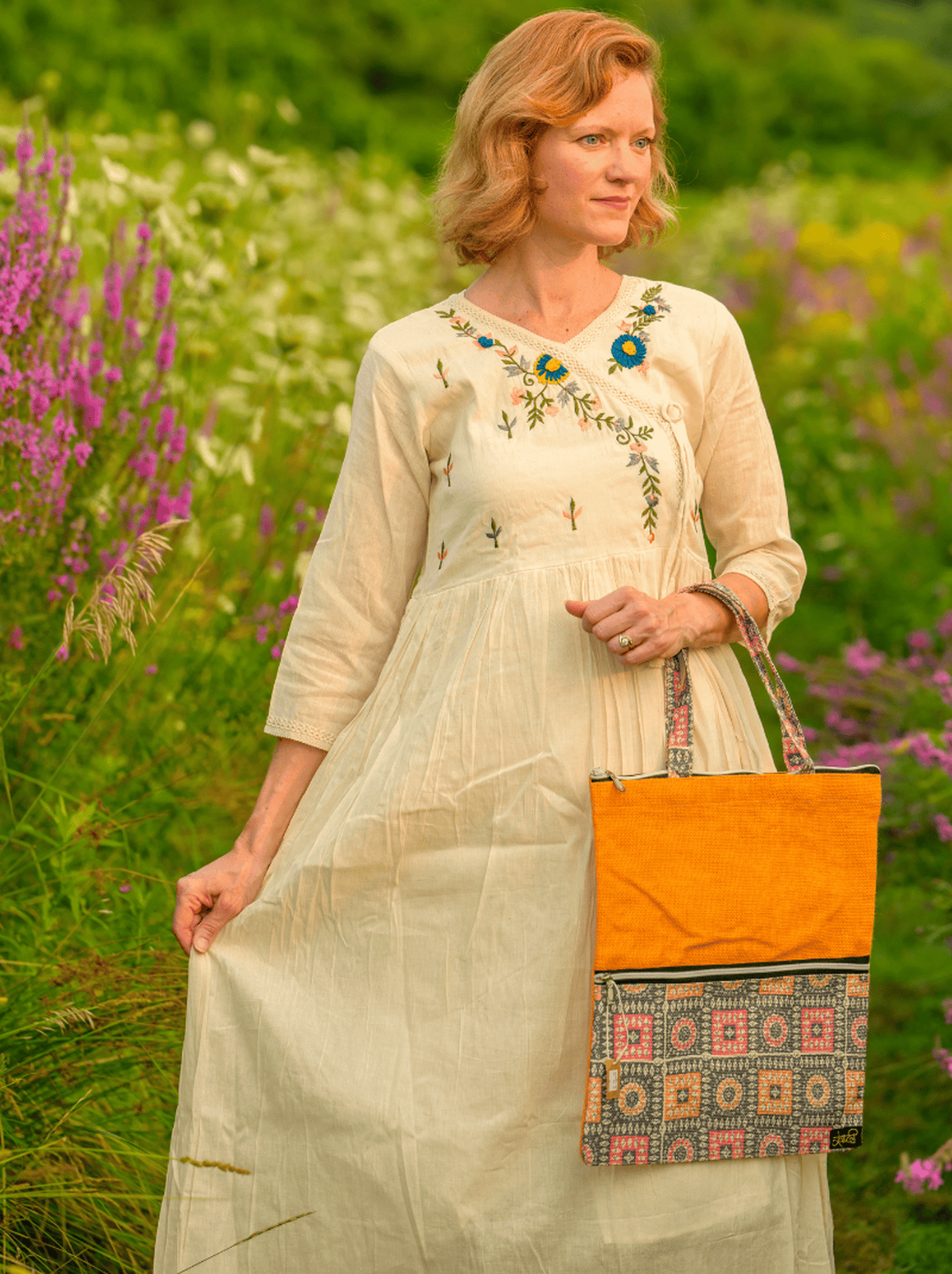 Woman in a light dress holding an orange bag with floral patterns in a field of flowers.