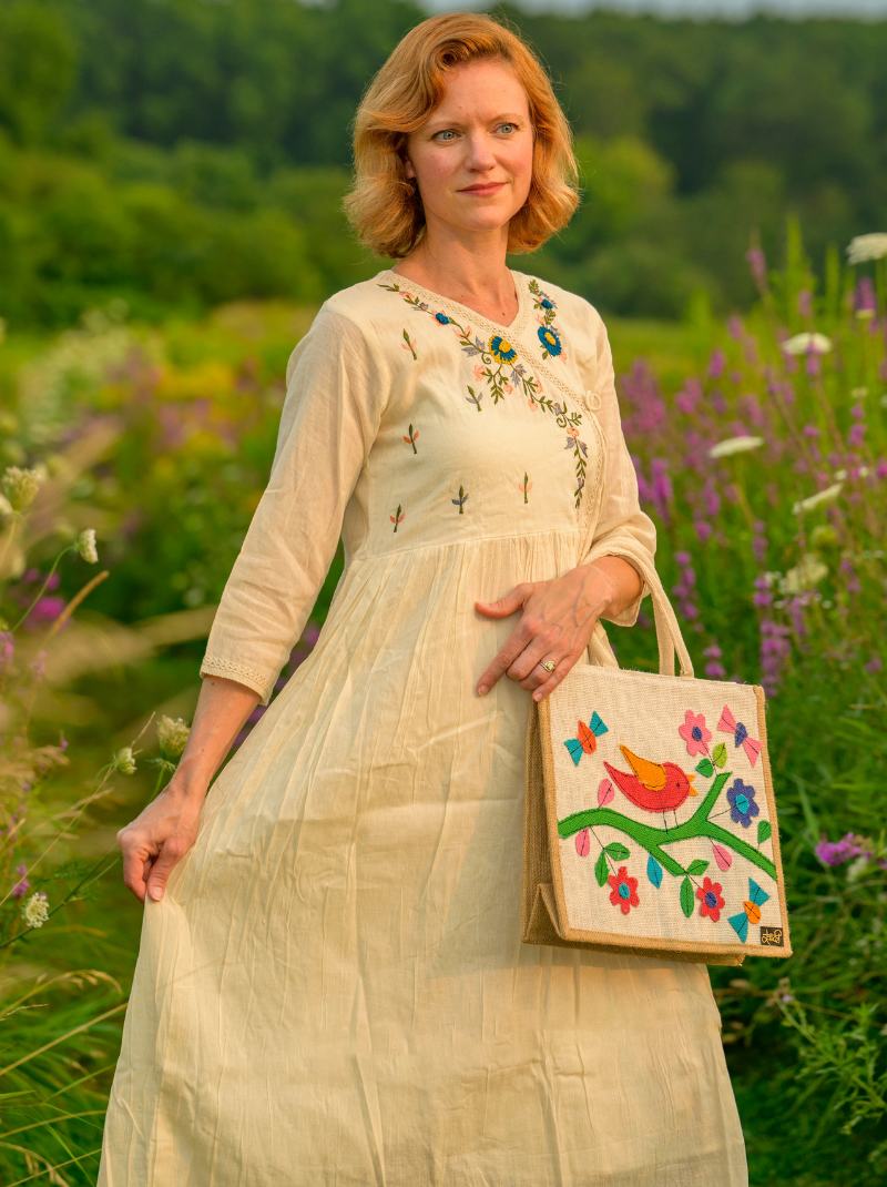 Woman in a beige embroidered dress holding a handbag with floral and bird design in a field.