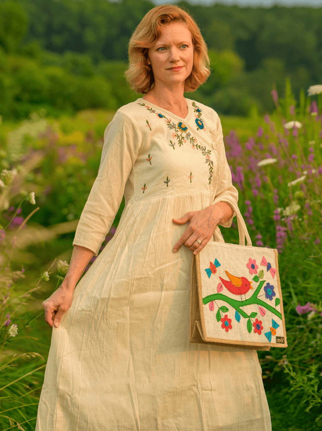 Woman in a beige embroidered dress holding a handbag with floral and bird design in a field.