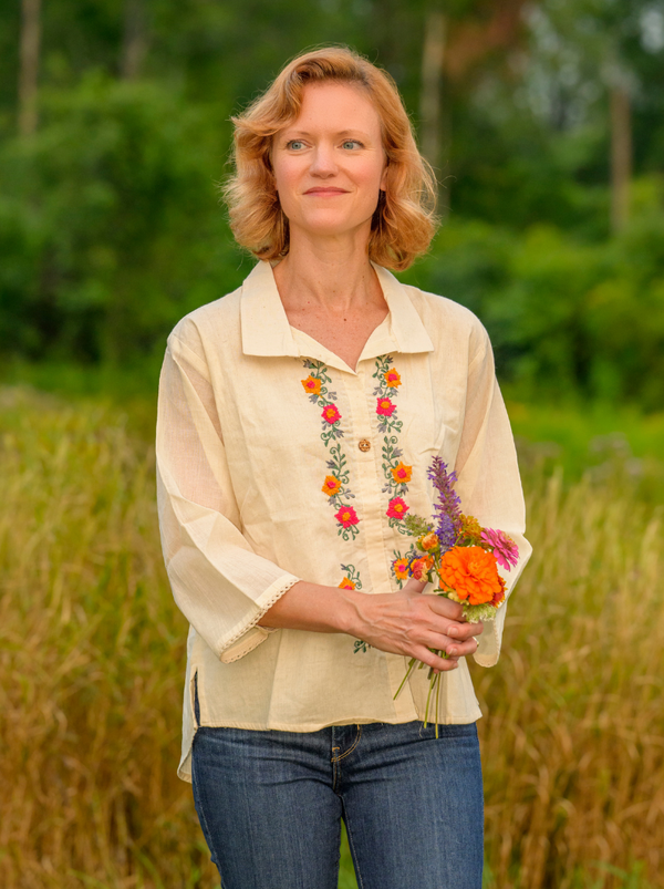 Woman wearing a beige blouse with floral embroidery, holding flowers, standing in a natural setting.