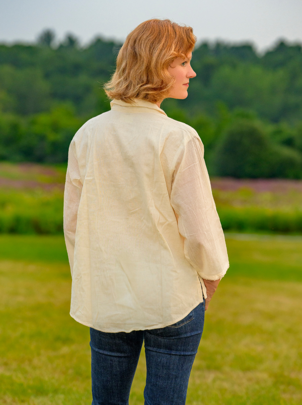 Woman standing in a field wearing a beige shirt and blue jeans, looking to the side.