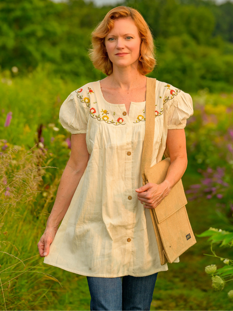 Woman wearing a beige embroidered blouse standing in a field with greenery and flowers.