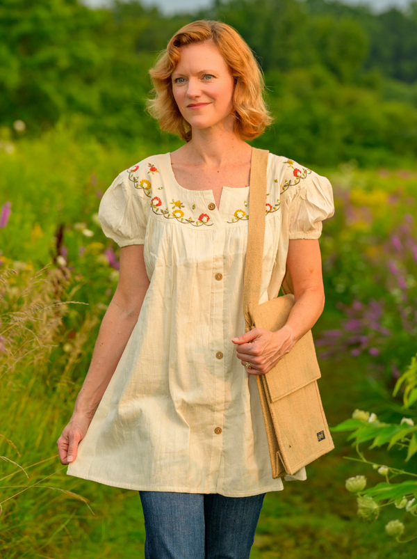 Woman in a light-colored embroidered dress standing in a field with greenery and flowers.