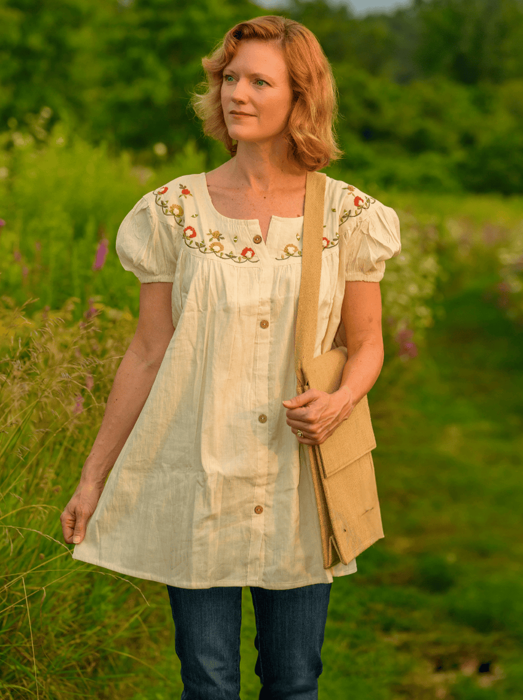 Woman in a light-colored embroidered dress standing in a field with greenery and flowers.