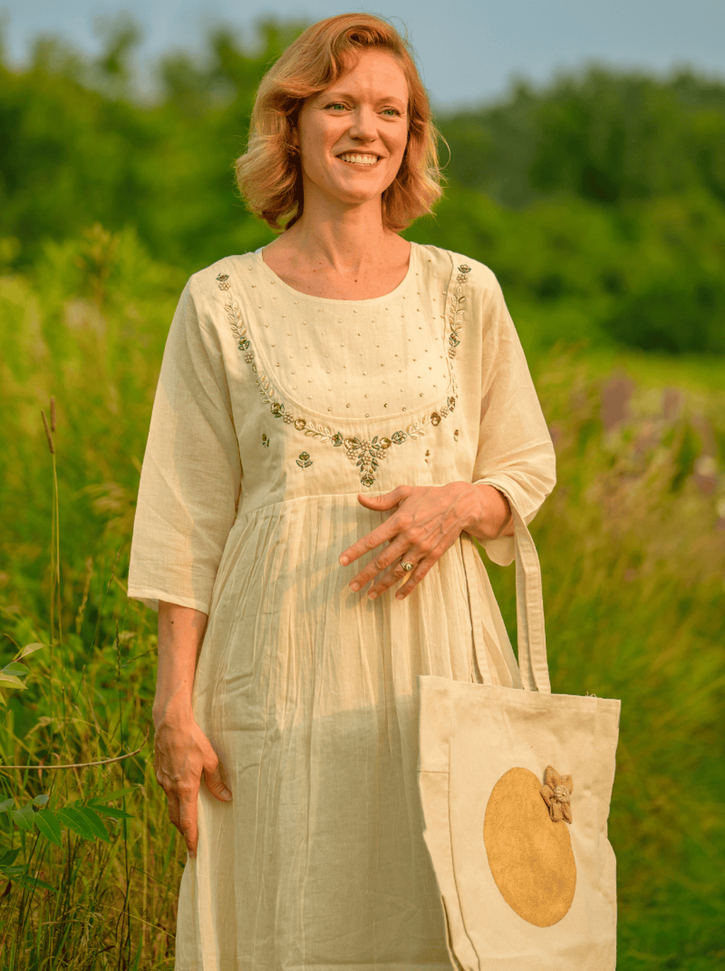 Woman in a light dress holding a tote bag with a floral pattern in a field.