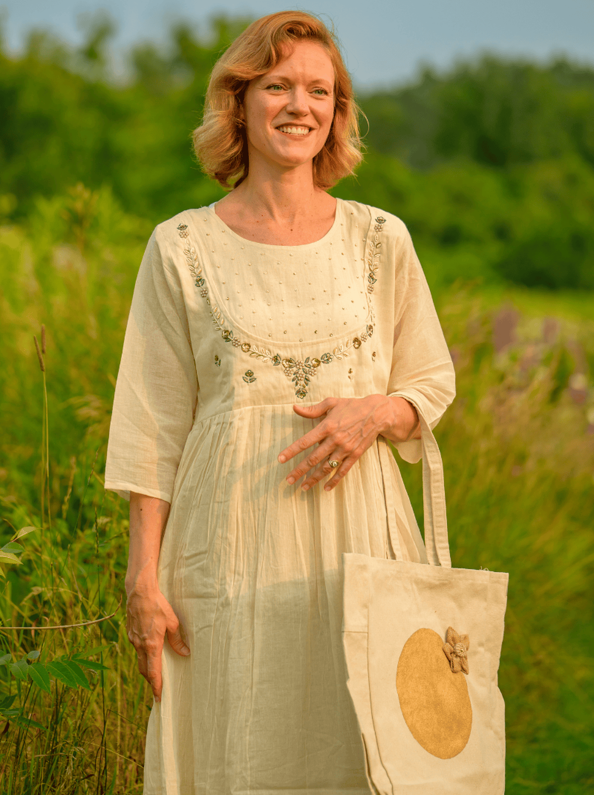 Woman in a light dress holding a tote bag with a floral pattern in a field.