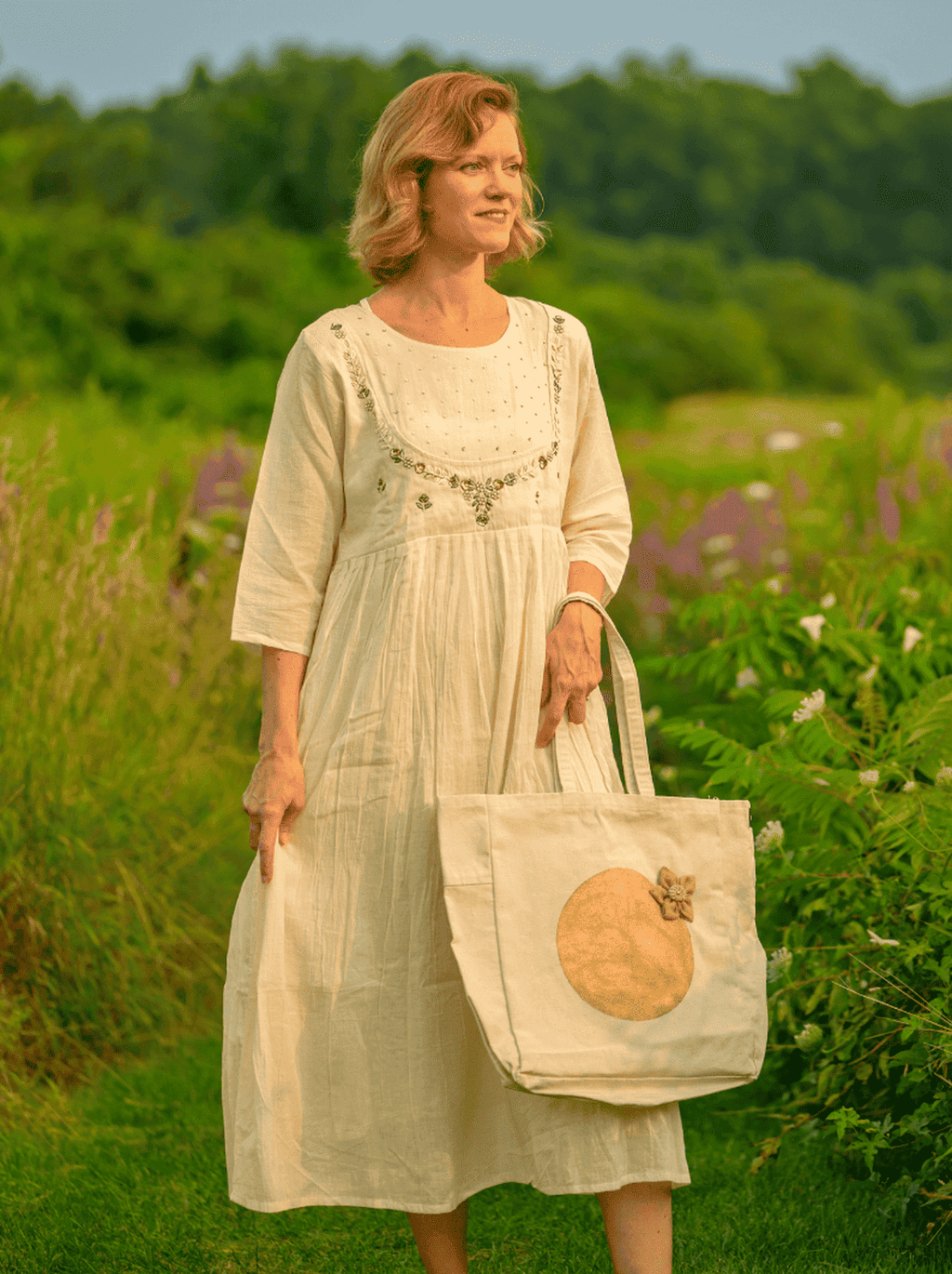 Woman in a light dress holding a tote bag with a pumpkin design in a field.