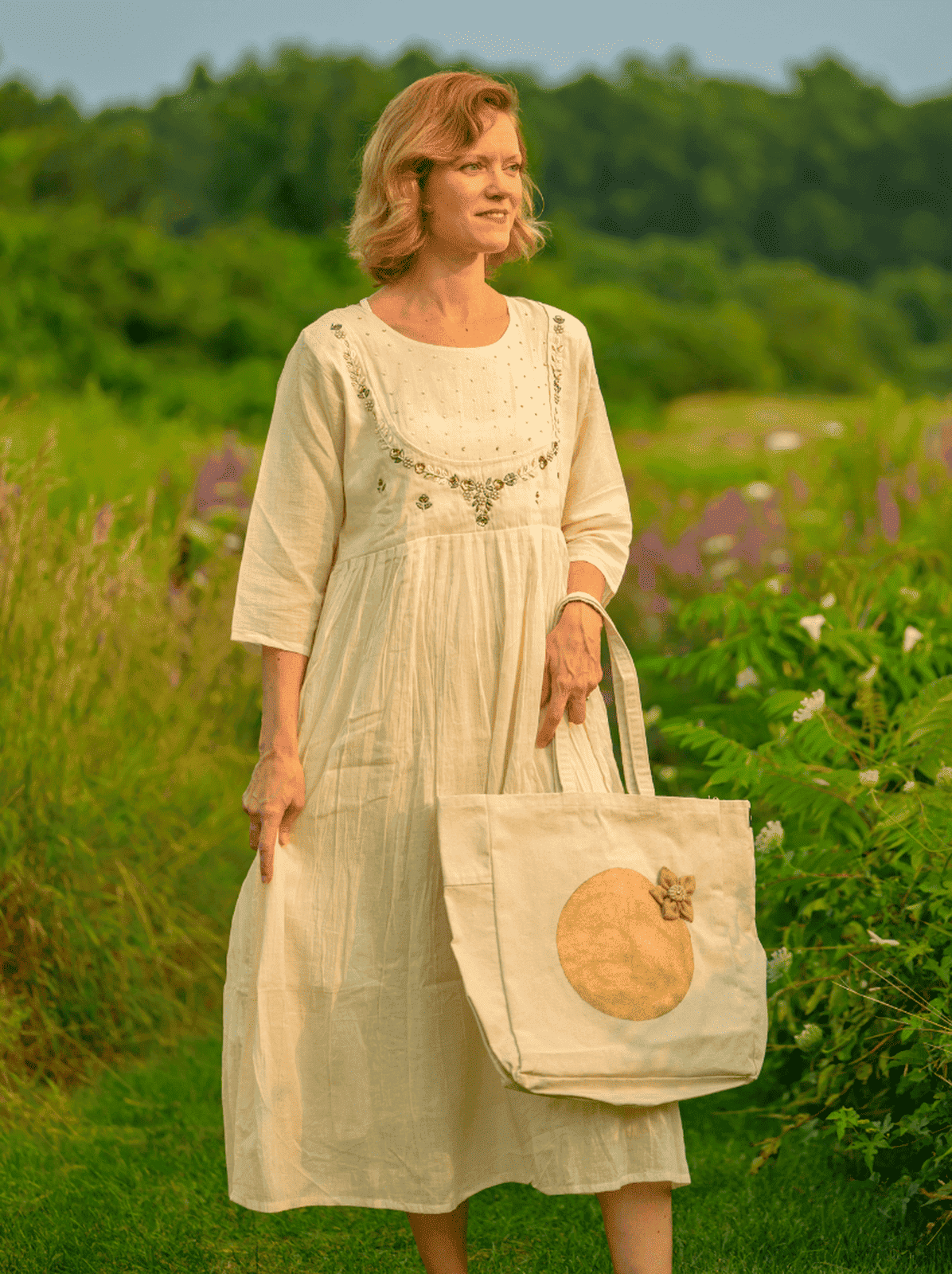 Woman in a light dress holding a tote bag with a pumpkin design in a field.