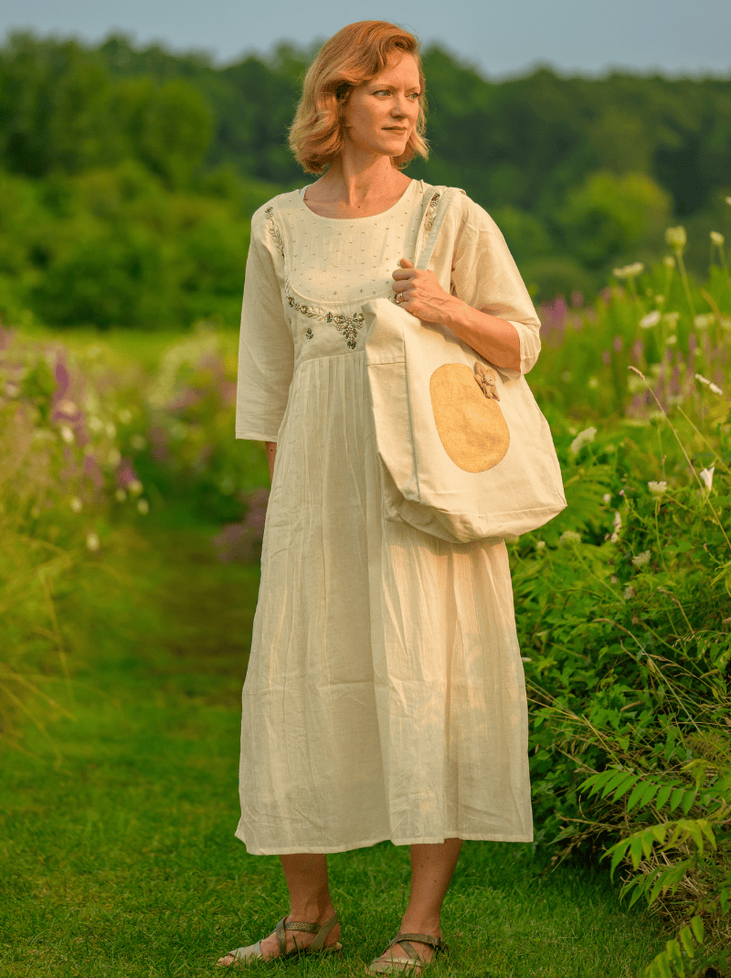 Woman in a long white dress holding a tote bag in a greenery and flowers.
