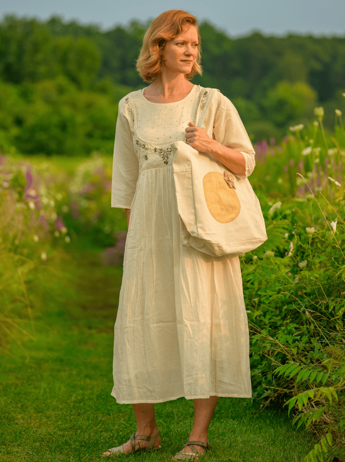 Woman in a long white dress holding a tote bag in a greenery and flowers.