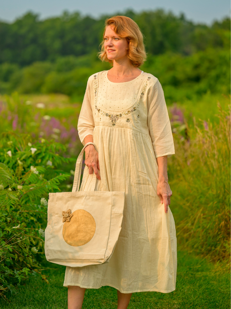 Woman in a white dress holding a tote bag in a field with greenery and flowers.
