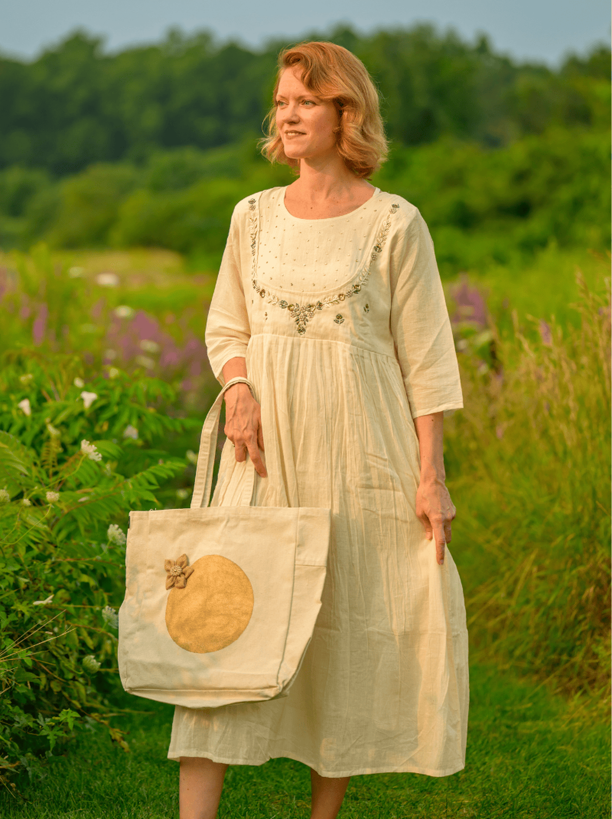 Woman in a white dress holding a tote bag in a field with greenery and flowers.