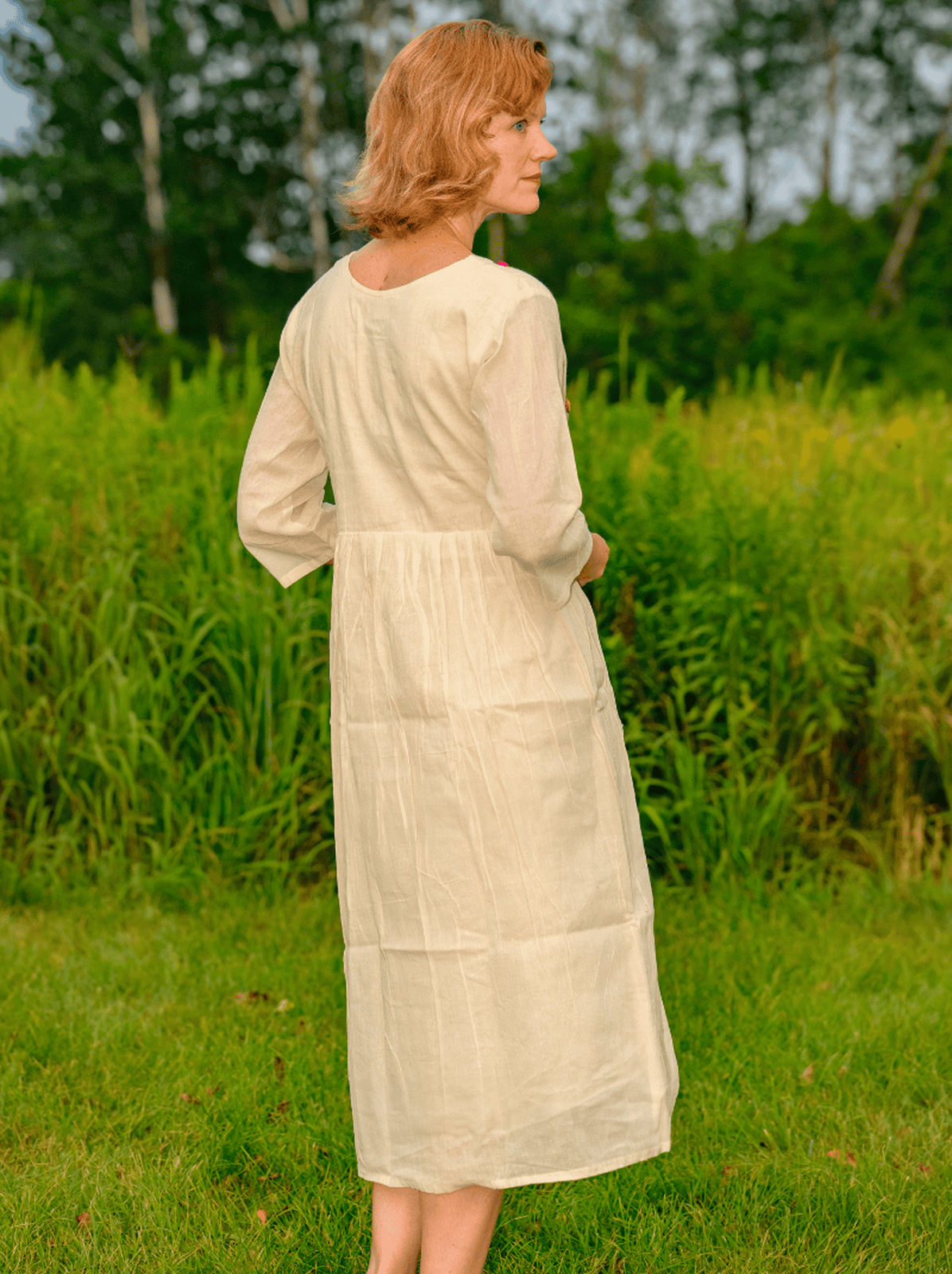 Woman in a long beige dress standing in a grassy field with trees in the background