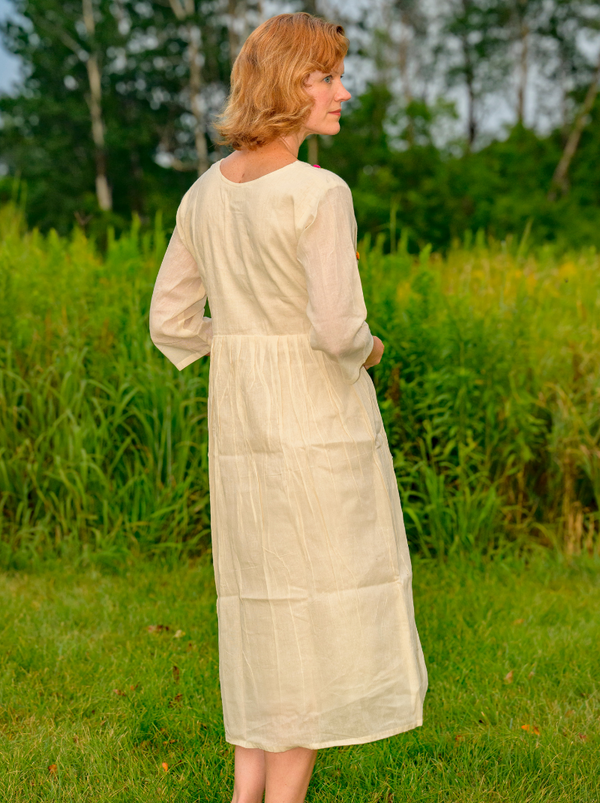 Woman in a long beige dress standing in a grassy field with trees in the background