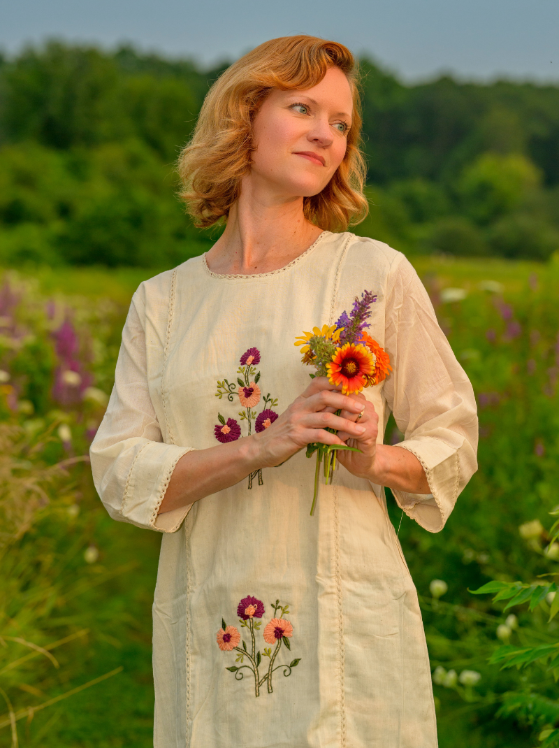 Woman in a white dress with floral embroidery holding a bouquet of flowers in a field.