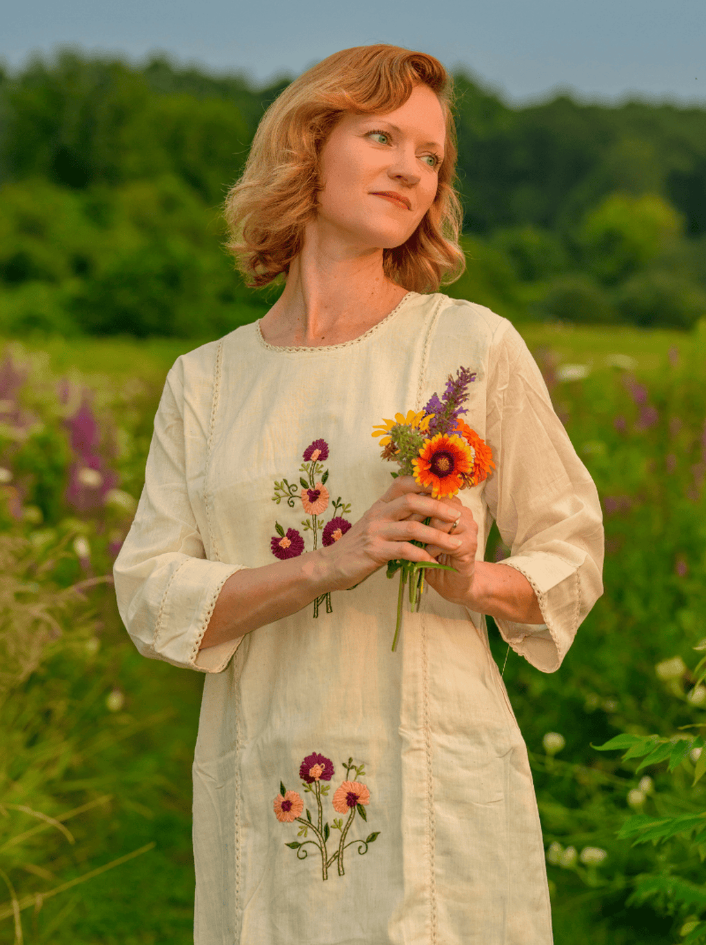 Woman in a white dress with floral embroidery holding a bouquet of flowers in a field.