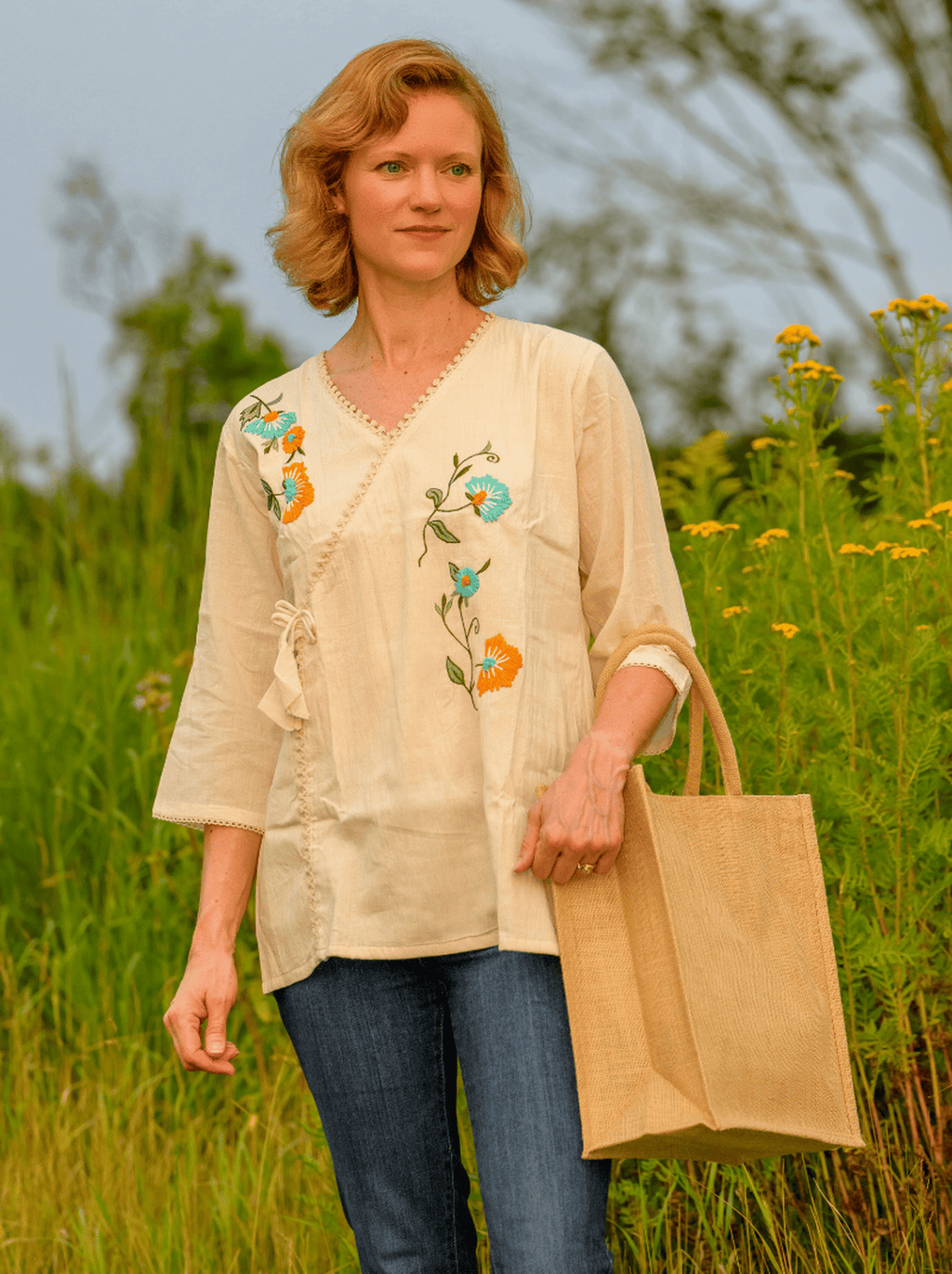 Woman in a beige embroidered blouse holding a brown paper bag in a field.