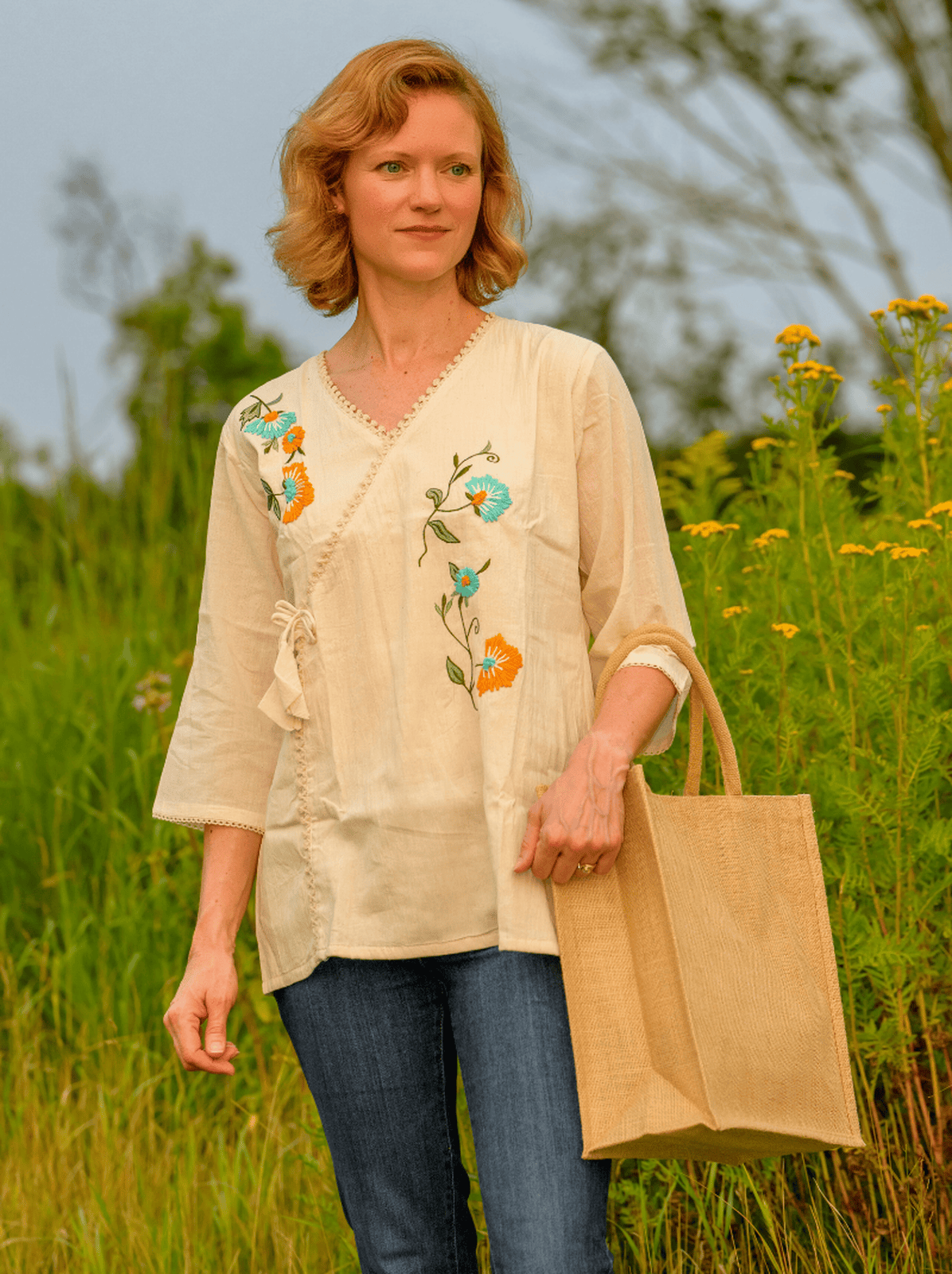 Woman in a beige embroidered blouse holding a brown paper bag in a field.
