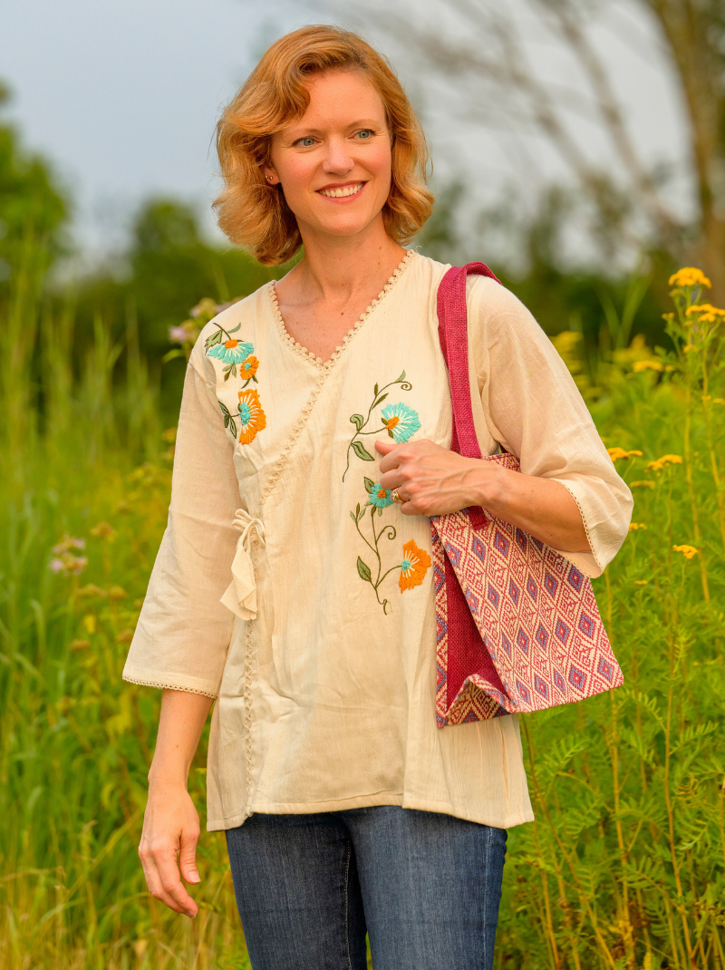 Woman in a beige embroidered top and jeans standing in a field with a colorful bag.