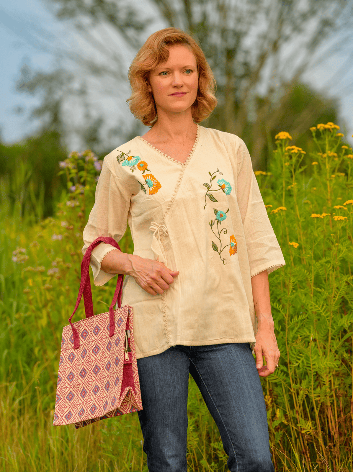 Woman in a beige embroidered blouse standing in a field with a pink bag.