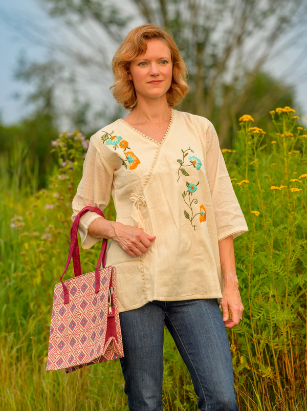 Woman in a beige embroidered blouse standing in a field with a pink bag.