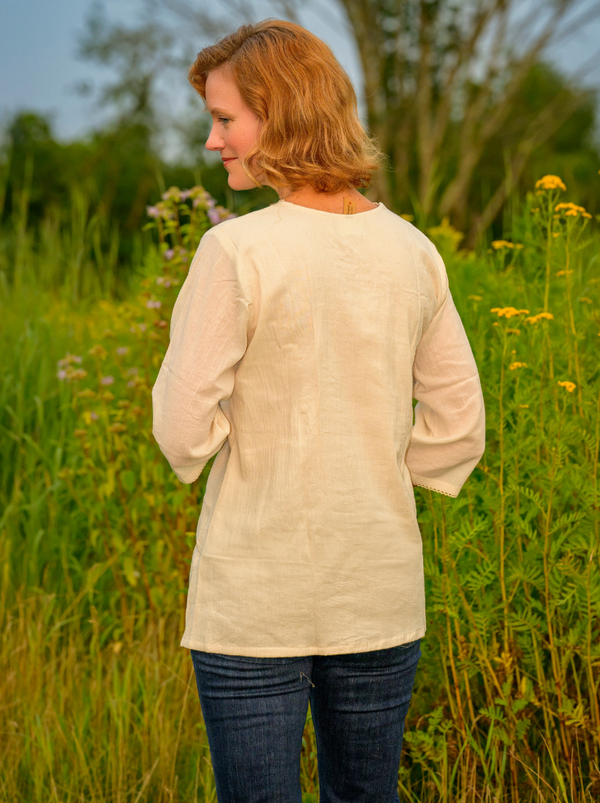 Woman wearing a beige long-sleeve shirt standing in a field with greenery and flowers.